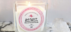 A lit white candle in a clear square glass container sits next to a cluster of crystals. The candle label reads Jade Orchid & Lotus Blossom Natural Scented Candle.