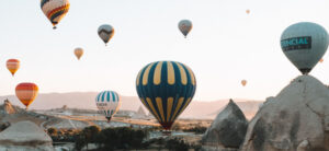 Hot air balloons of various colors and patterns floating in the early morning sky over a rocky landscape, with sunlight illuminating the scene.