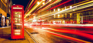 A red telephone booth stands on a bustling city street at night. Bright, colorful streaks of light from passing traffic illuminate the scene, creating dynamic motion effects against the backdrop of lit buildings.