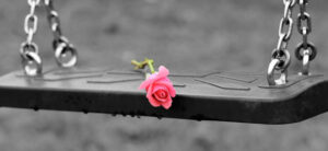 A single pink rose lies on a black rubber swing seat, with gray, blurred background, creating a somber yet tender contrast.
