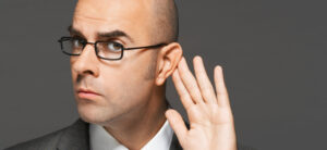 A man with glasses, wearing a suit, holds his hand to his ear, appearing to listen closely. The background is a solid gray color.