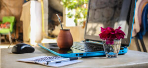 A laptop with a blue edge sits open on a table next to a mouse, a notepad, and a pen. A wooden mate cup and a glass of pink flowers are also on the table. The background shows a blurred outdoor scene.