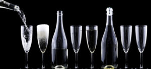 A row of seven champagne flutes on a black background. The first glass is being filled with bubbly liquid, the second is full with foam, and the others are empty. Two champagne bottles are placed among the glasses.