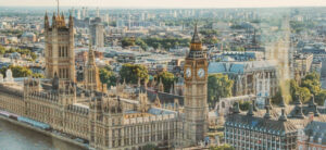 Aerial view of the Palace of Westminster in London, featuring the Elizabeth Tower with the Big Ben clock. The Thames River is visible to the left, with urban buildings and greenery in the background under a clear sky.