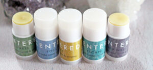 Five small cylindrical containers with different colored labels: green, blue, yellow, teal, and gray. Each labelled with a word: DE-STRESS, FOCUS, HAPPY, ESCAPE, and SLEEP WELL for applying to various points like wrists and temples.