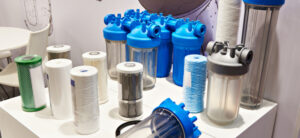 Various water filter cartridges and housings displayed on a table. The filters vary in size and design, with some encased in transparent housings and others in blue and gray tops. Coiled and cylindrical filter elements are also present.