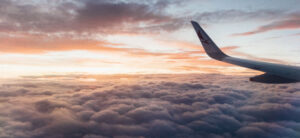 View from an airplane window showing a wing against a backdrop of fluffy clouds and a colorful sky at sunset. The horizon displays shades of orange, pink, and blue as the sun sets.