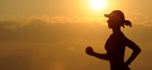 Silhouette of a person jogging at sunrise or sunset, with a bright sun in the background. They are wearing a sports cap and athletic clothing, with a serene sky and a few clouds.