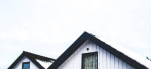 Two white wooden house rooftops with dark trim, partially covered in snow, against a cloudy sky background. The foreground house has a single window beneath the roof peak.