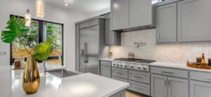 Modern kitchen with sleek gray cabinets and stainless steel appliances. Features a white countertop with a gold vase holding green leaves. A gas stove is under a vent hood, and a patterned backsplash adds detail. Large window provides natural light.