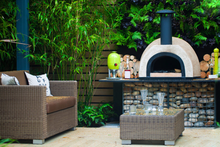 A cozy outdoor patio area with a wicker sofa, a small table topped with glassware, and a stone pizza oven surrounded by stacked logs and lush green plants in the background.