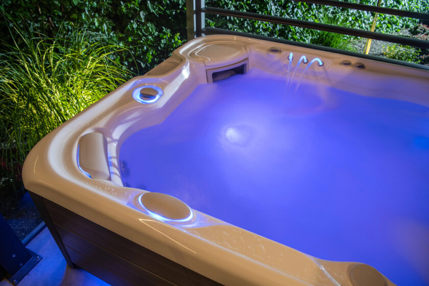 A hot tub filled with glowing blue water at night, with jets creating gentle streams. The tub is surrounded by greenery and illuminated by soft, ambient lighting.