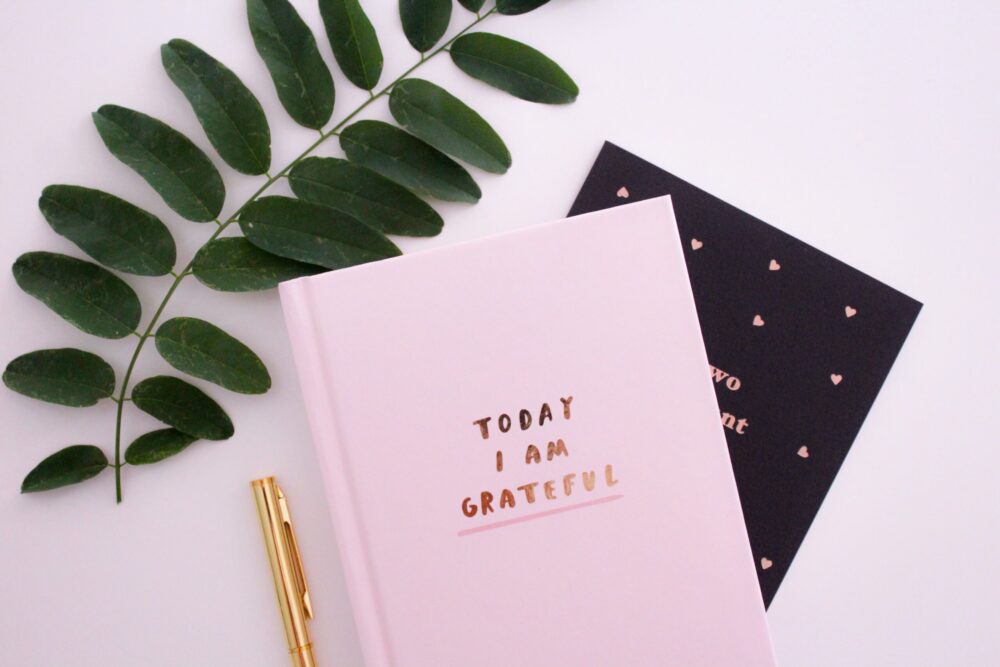 A pink notebook which has the worlds Today I am Grateful on the Front, sat on top of a black and pink notebook, a gold pen to the left side and some leaves to the top
