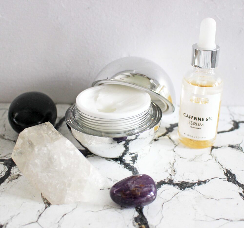 skinchemists products the Anti-Ageing Caviar Night Moisturiser on the right, it is in a silver reflective round tub the lid off and leaning on the side, on the left the bi-phase caffeine serum bottle which contains a yellow liquid