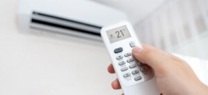 A person adjusts an air conditioning unit using a remote control. The remote display shows a temperature setting of 21°C. The air conditioner is mounted on a wall in a bright room.