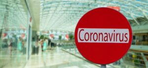 Red circular sign with the word Coronavirus in white letters is shown in the foreground. The background features a blurred view of a spacious, glass-roofed shopping mall with people walking inside.
