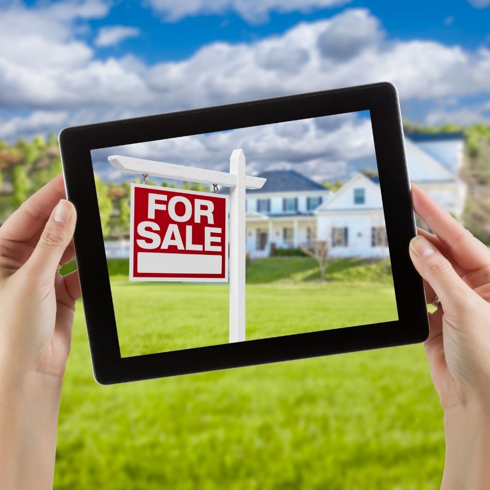 person holding a black frame around a for sale sign with houses in the background