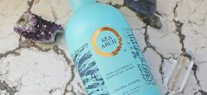 A bottle of Sea Arch non-alcoholic distilled spirit with a turquoise label featuring a gold circular logo and text. It is placed on a cracked surface, surrounded by amethyst crystals and a clear quartz.