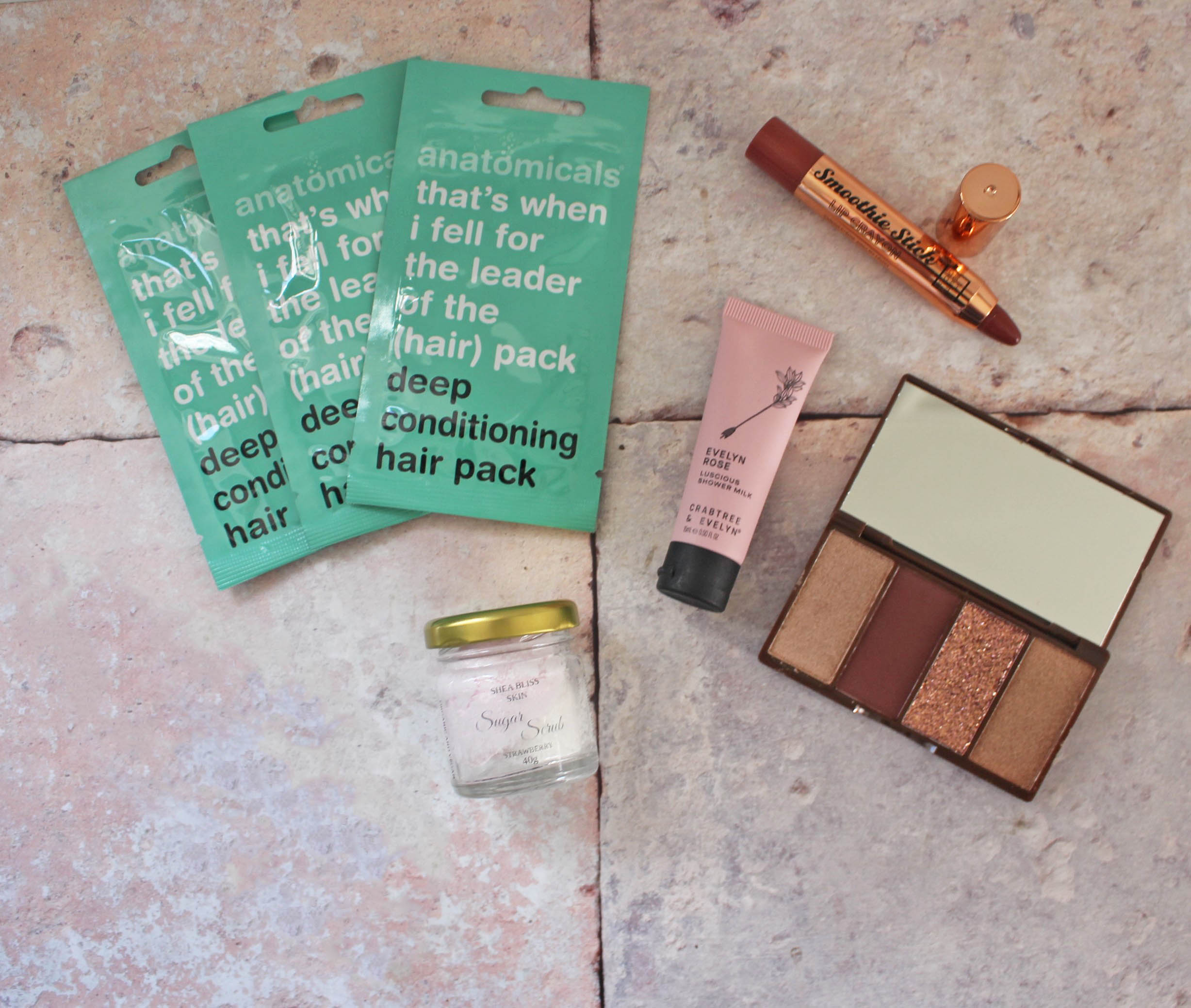 Flat lay of beauty products on a textured surface: three green hair pack sachets, a small jar with a yellow lid, a pink tube, a lipstick, and an open palette with three shimmery shades.
