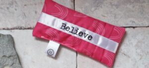 A small rectangular pink pouch with a white ribbon across it, featuring the word “Believe” in black text. The fabric has a subtle wave pattern. A small tag with a circular design is attached at the bottom. The pouch rests on a stone surface.