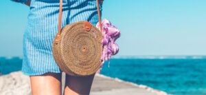 someone walking alongside the sea wearing a blue dress and using a woven bag