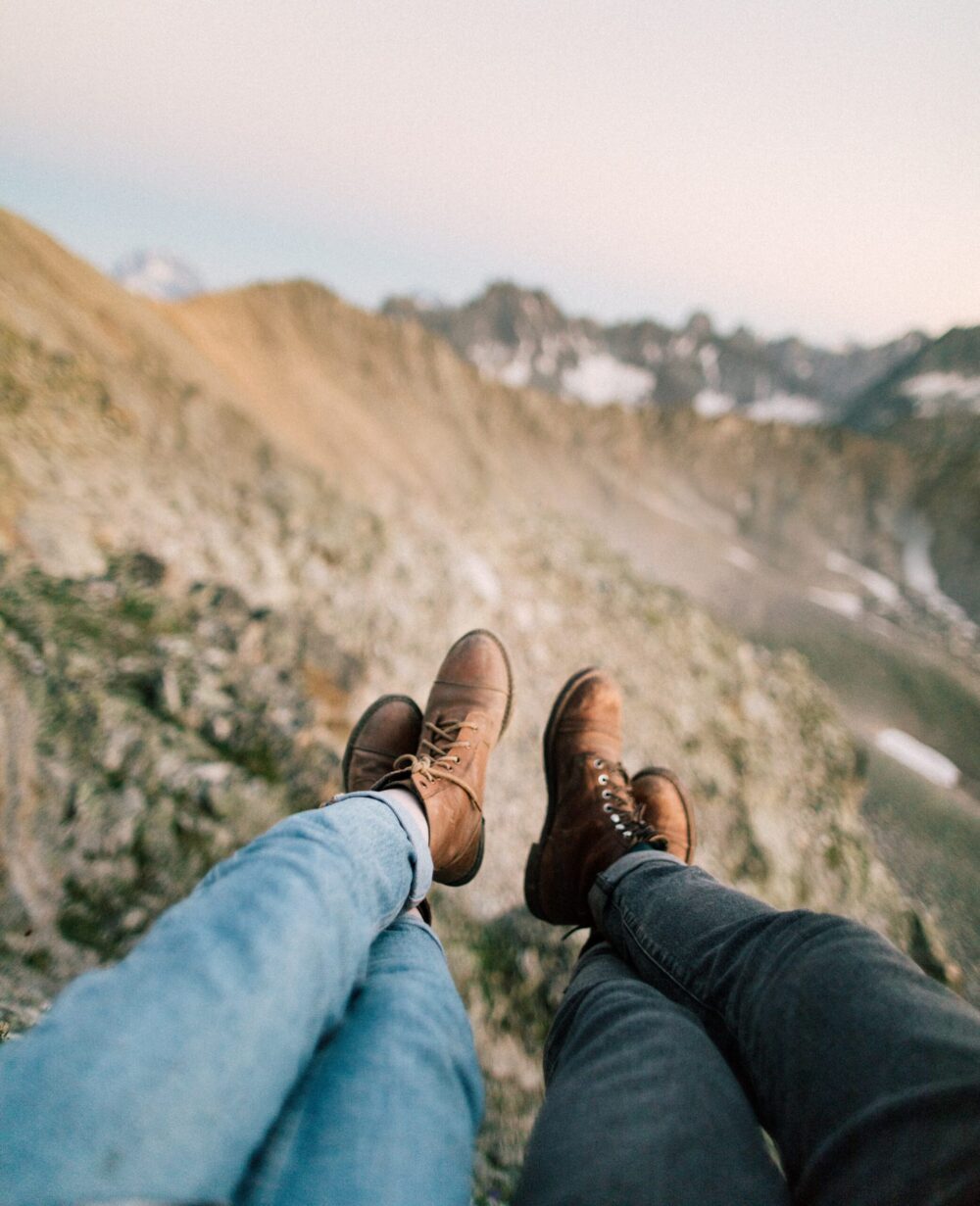 Two people sat on a mountain with their legs out in front of them wearing jeans and walking boots