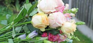 A bouquet of pale pink and cream roses, artfully arranged with LÖV Flowers signature touch of pink and lavender blooms, is complemented by lush green leaves, all set against a blurred garden background.