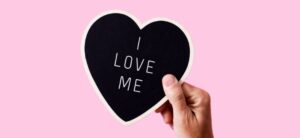person holding a heart that says I LOVE ME against a pink backdrop her