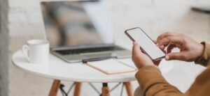 person holding a smart phone with a laptop in the background
