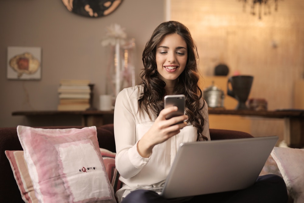 Woman sitting on a sofa while looking at phone and on her laptop