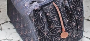 A navy blue bag adorned with geometric patterns and a rose gold zipper, complete with a round pull tab, rests on a textured white surface, ready to smash expectations with its striking design.