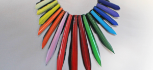 A Laura Zabo necklace crafted from multicolored elongated pencil-shaped beads arranged in a semi-circle. Each bead showcases a unique color, creating a vibrant and artistic rainbow effect against a light background.