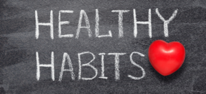 The image shows the phrase Healthy Habits written in white chalk on a blackboard. A red heart shape replaces the letter O in Habits, adding a visual emphasis on the theme of well-being.
