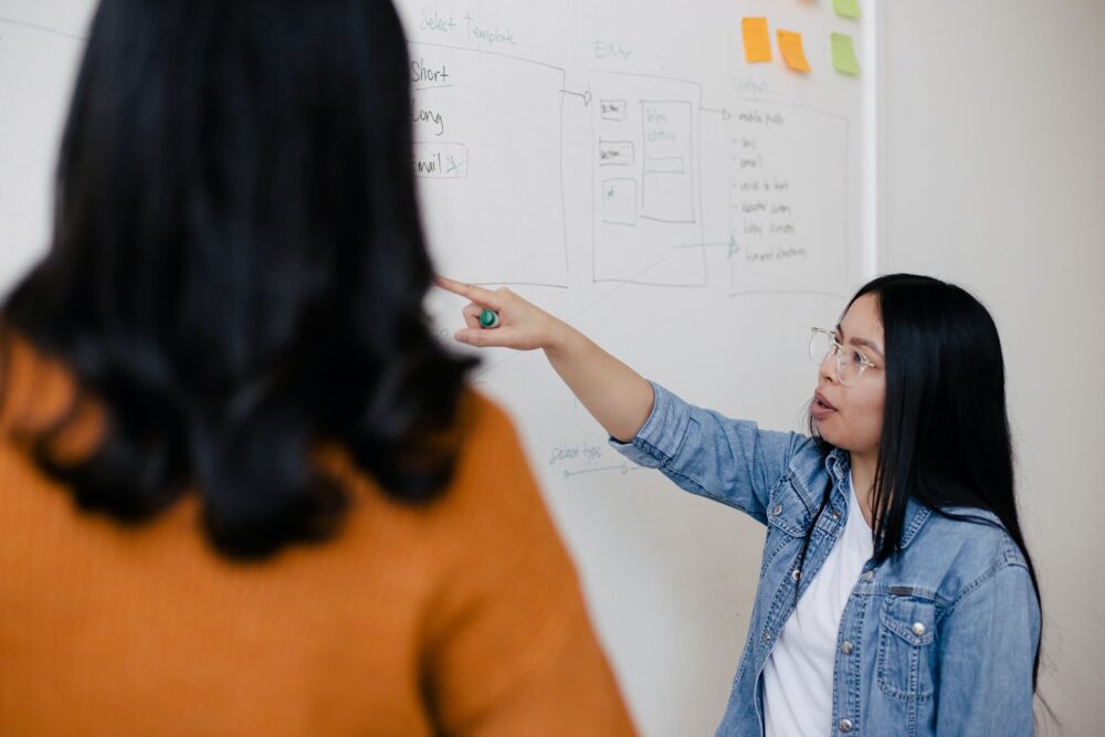 two women standing in front of a white dry erase board