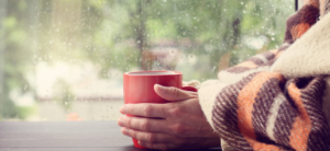 person sat a window wrapped in a blanket with hands around a red mug