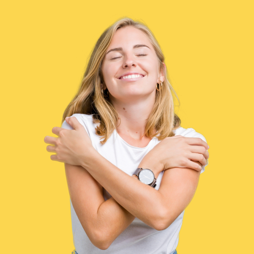 woman with blonde hair against a yellow backdrop hugging herself
