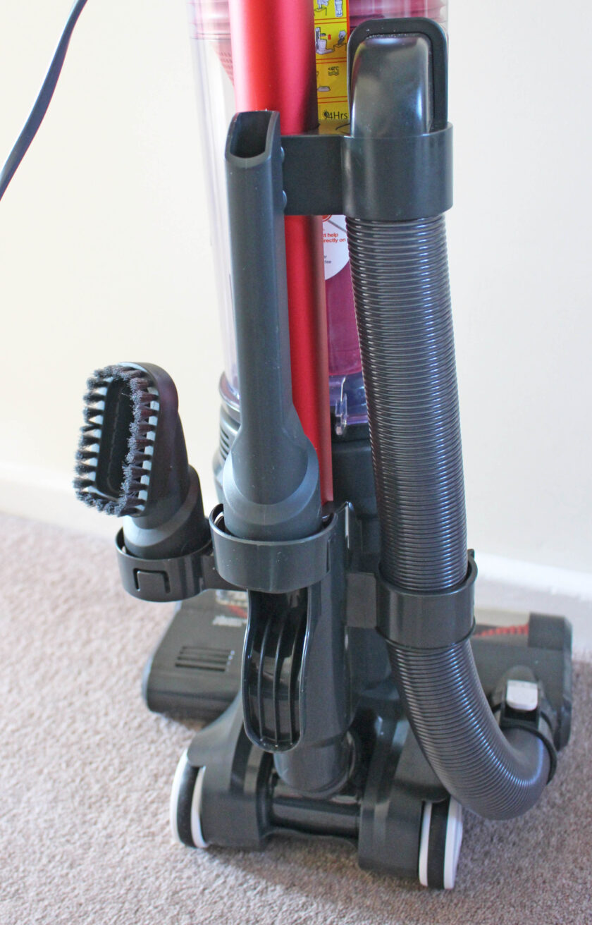 A close-up view of the Upright 300 Vacuum Cleaner on a beige carpet. This red vacuum features various attachments, including a bristle brush and a nozzle, secured on its side. A flexible hose is also visible, coiled neatly around the unit.