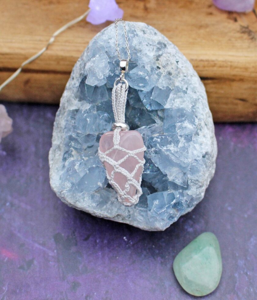 A rose quartz pendant with silver wire wrapping rests on a large, rough blue crystal. A small green stone is nearby, all set against a purple and wooden surface, delicately tied together with good twine for an artistic touch.