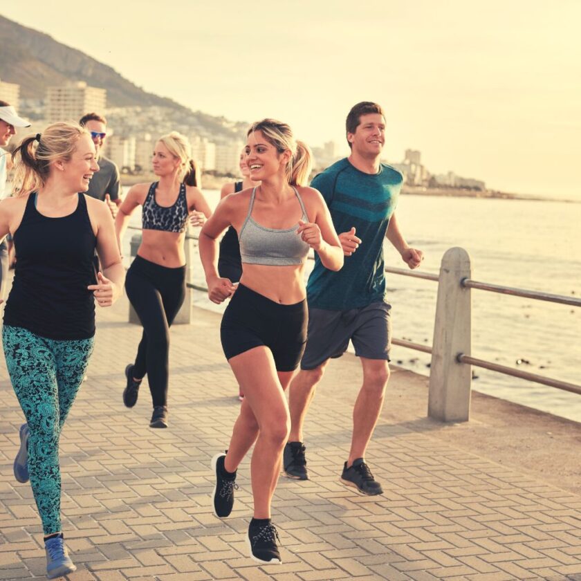 A group of people jogs along a seaside promenade. They are wearing athletic clothing. The sun is setting, casting a warm glow, and buildings and mountains are visible in the background.