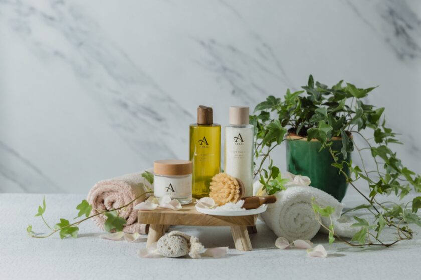 A wooden tray with ARRAN skincare products, including bottles and jars, is surrounded by rolled towels, a brush, and a green plant. Petals are scattered on a white marble-like surface, enhancing the serene spa-like setting.