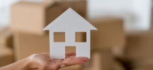person holding a small white cut out house with packing boxes in the background