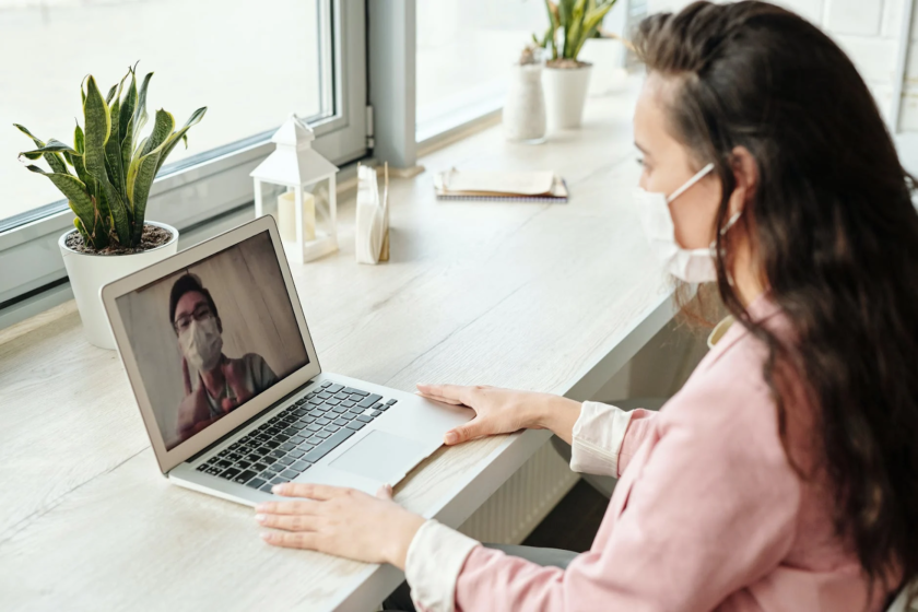 A woman in a pink shirt sits at a desk in front of a laptop, participating in a video call. Both she and the person on the screen are wearing face masks. The desk is by a window, with plants and decorative items nearby.