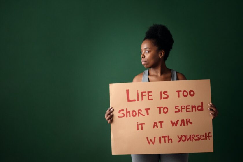 Woman holding a cardboard with “life is too short to spend it at war with yourself” written on it.