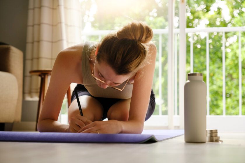 Young caucasian woman making a journal entry while sitting on a yoga mat at home with water bottle standing next to mat. Health and wellness concept.
