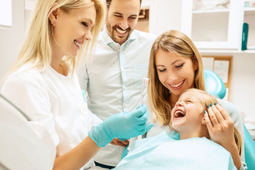 A dental hygienist holding a toothbrush smiles at a laughing child in a dental chair. The child is seated on a womans lap, who is also smiling. A man stands behind them, looking on with a cheerful expression. The setting is a dental office.