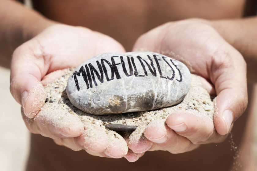 closeup of a young caucasian man, on the beach, with a handful of sand in his hands and a stone on top with the text mindfulness written in it