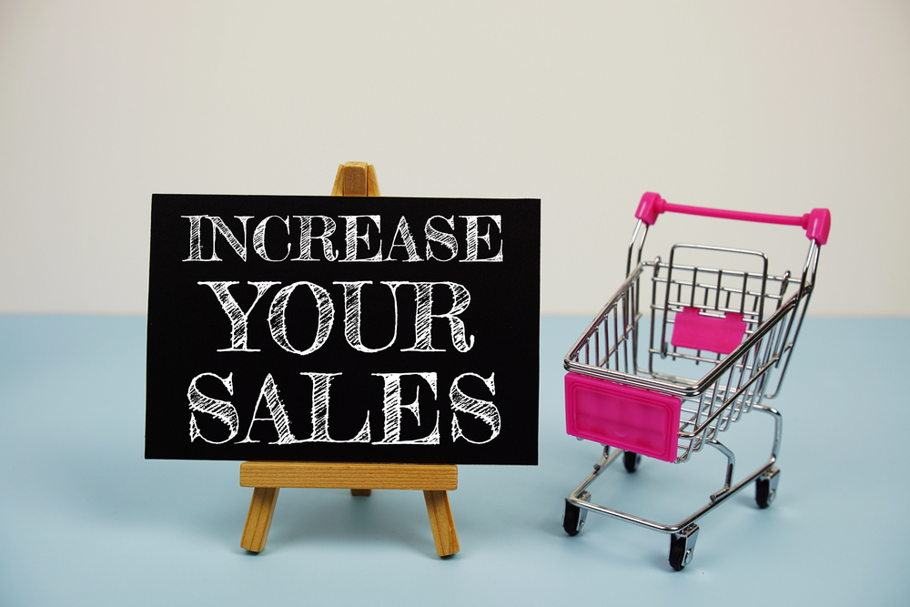 A small wooden easel displaying a sign that reads INCREASE YOUR SALES, next to a miniature pink shopping trolley.