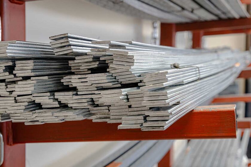 A stack of silver metal bars neatly arranged on red metal shelves in a warehouse-style setting. The bars are rectangular and reflect light, emphasizing their metallic surface.