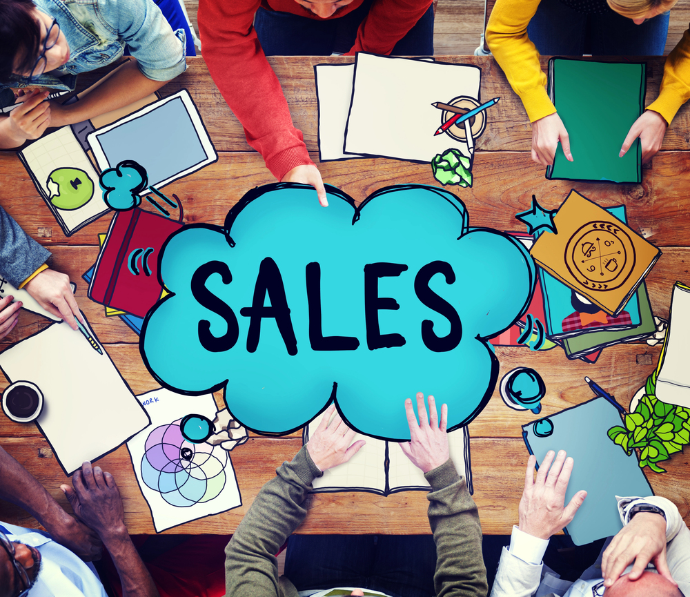 Overhead view of a group of people sitting around a wooden table cluttered with papers and tablets, surrounding a large teal speech bubble that says SALES in the centre.