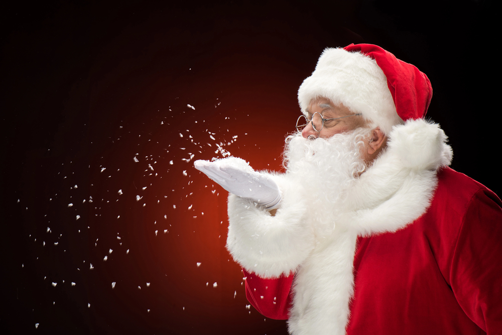 Father Christmas in a red suit and hat blows snowflakes from his gloved hand against a dark background with a warm orange glow.